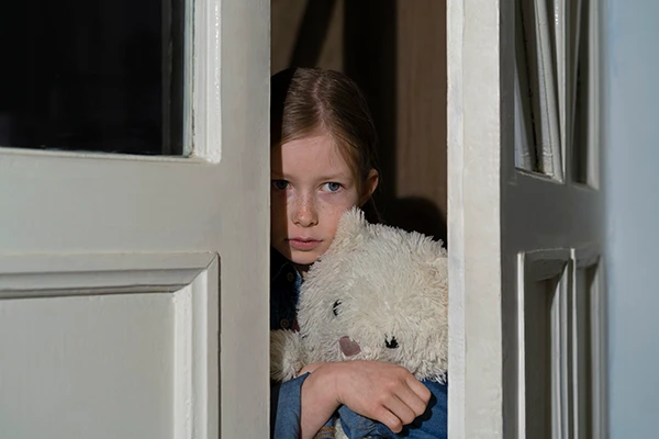 Anxious child holding a teddy bear for comfort, peering out from behind a cracked closet door.