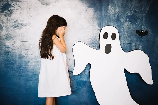 Scared young girl covering her face beside a ghost cutout, depicting childhood fears and anxiety.