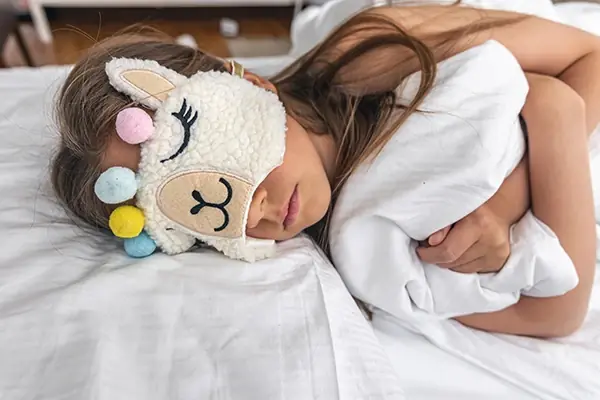 Young girl sleeping in bed wearing a plush llama eye mask to help manage bedtime anxiety and promote rest.