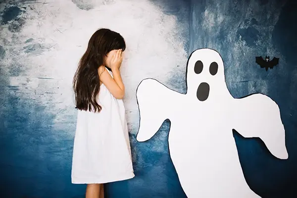 Scared young girl covering her face beside a ghost cutout, depicting childhood fears and anxiety.